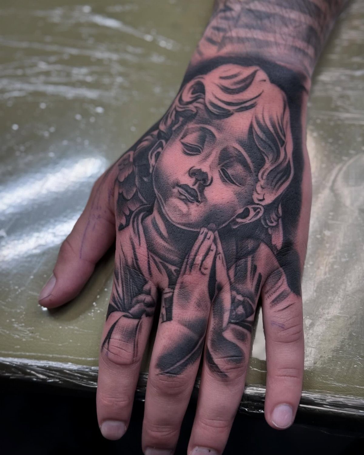 Detailed black and grey tattoo of a praying angel on a person's hand.