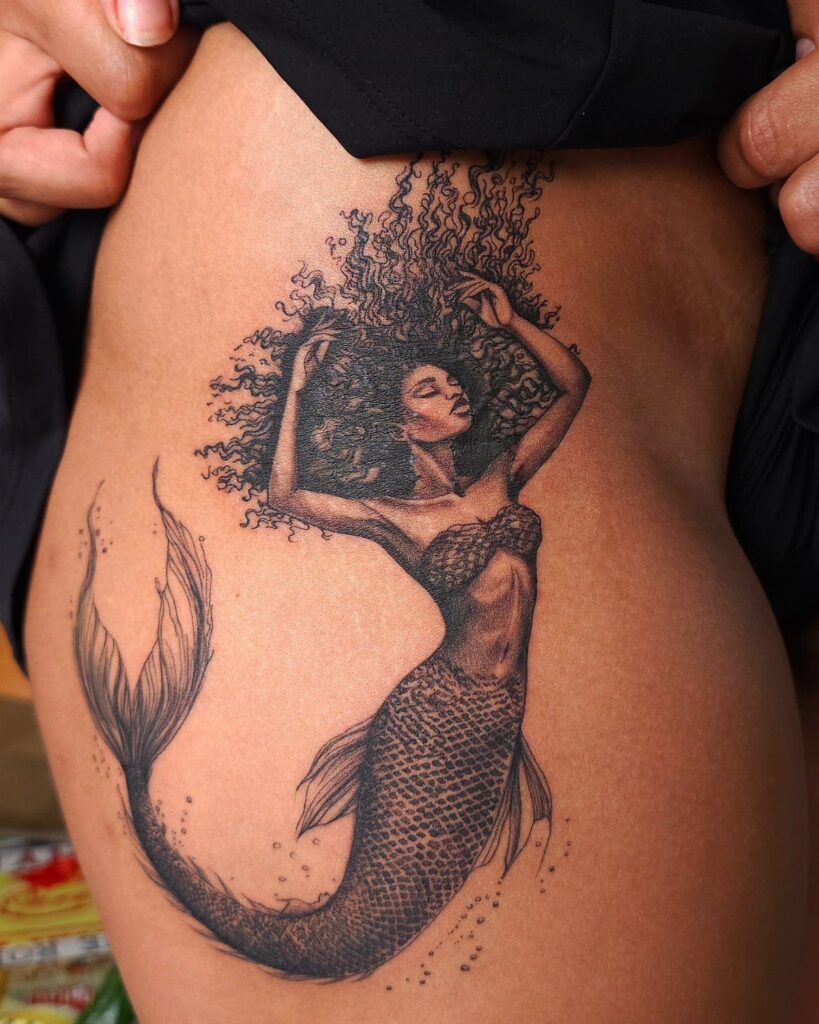 Black ink mermaid tattoo with curly hair and scaled tail on a person's hip