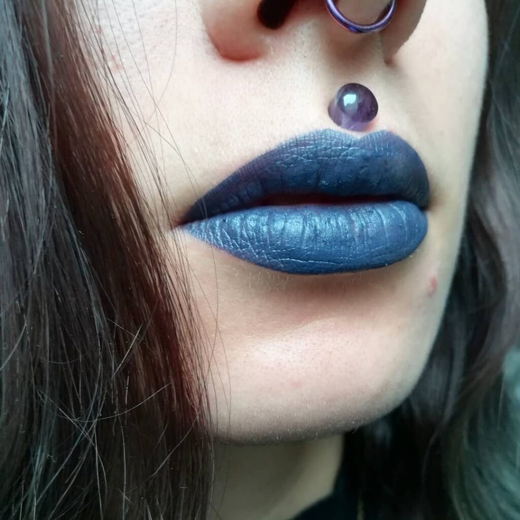 Person with blue lipstick and a purple bead Medusa piercing above the lip.