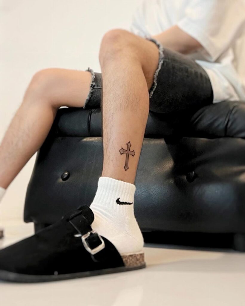 Small black cross tattoo on a person's lower leg and shin.