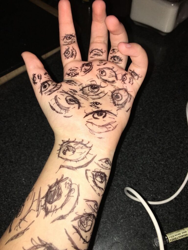 Hand and wrist covered in black ink drawings of multiple stylized eyes.