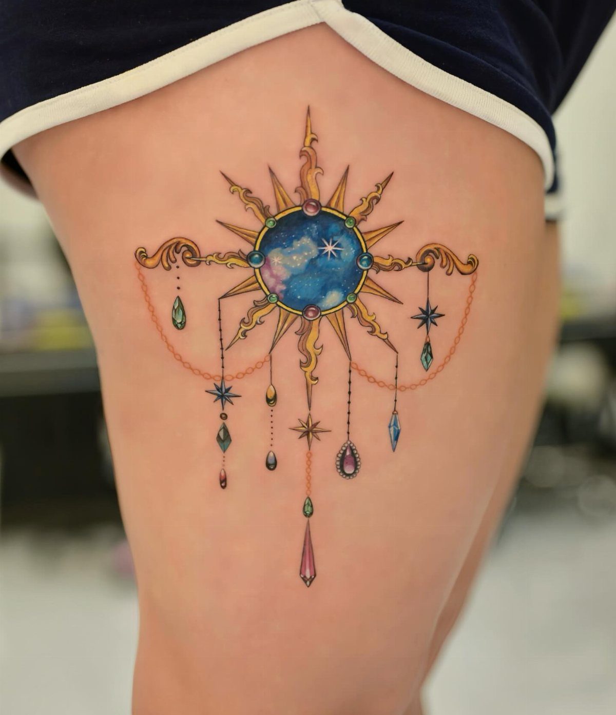 Golden sunburst tattoo with a blue cosmic center and hanging jewels on a thigh
