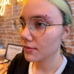 Person with green hair wearing glasses and lip and eyebrow piercings.