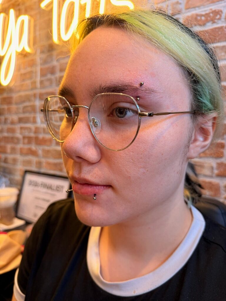 Person with green hair wearing glasses and lip and eyebrow piercings.