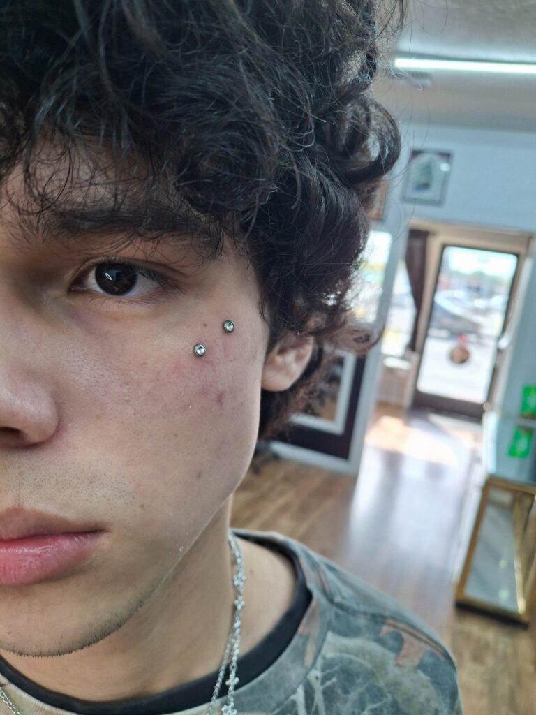Young man with curly hair showing two silver stud piercings on his cheekbone.