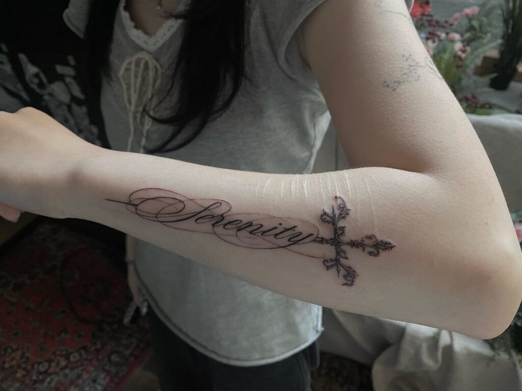 Forearm tattoo with the word Serenity in cursive and a decorative cross.