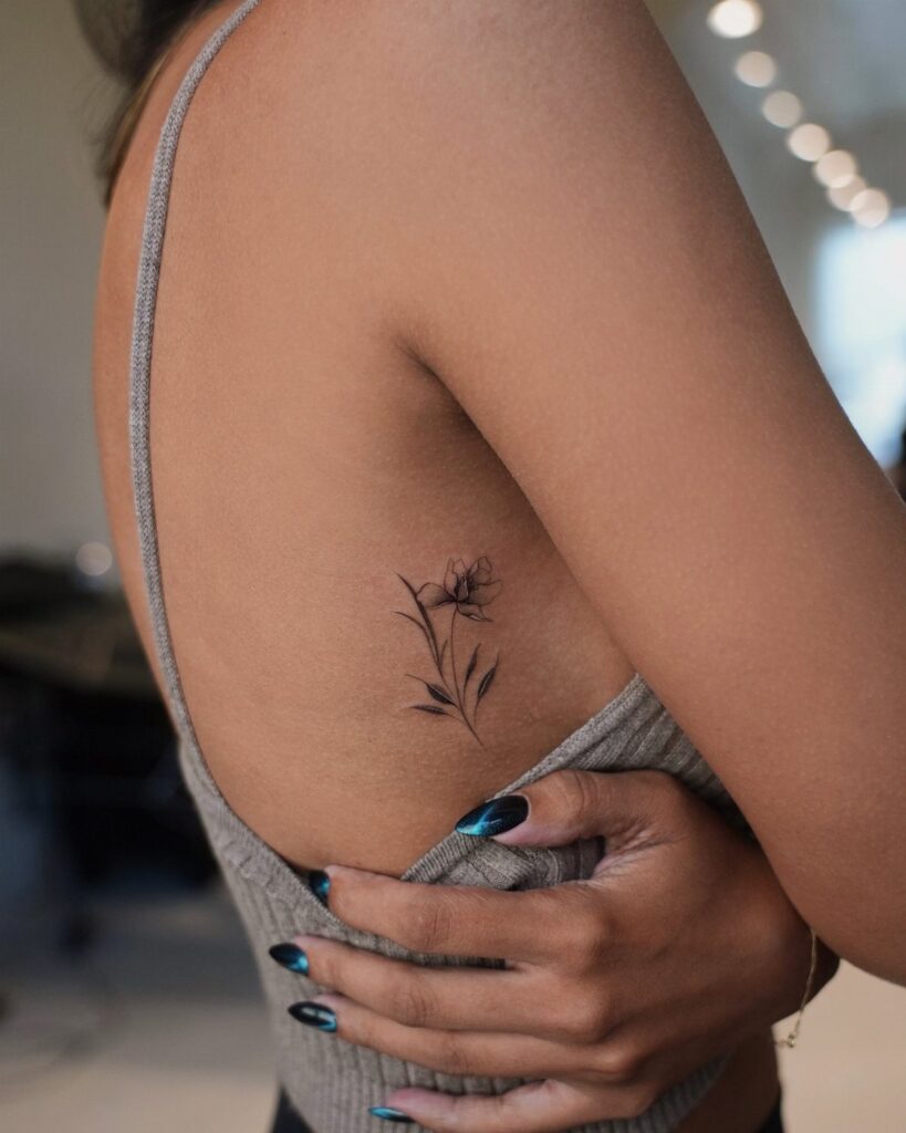 Floral black ink tattoo on the ribcage of a woman wearing a grey top.