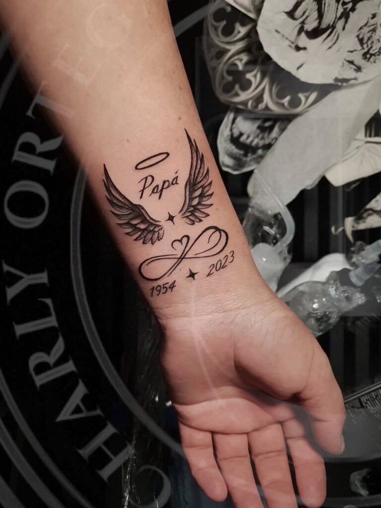 Memorial tattoo on a wrist with angel wings, halo, and dates for a father.