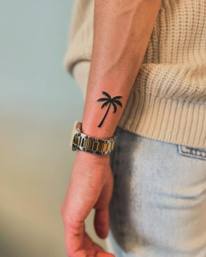 Small black palm tree tattoo on a person's wrist next to a metal watch.
