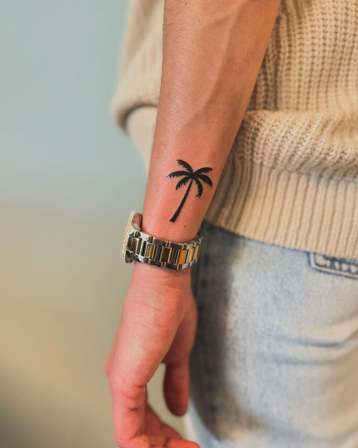 Small black palm tree tattoo on a person's wrist next to a metal watch.