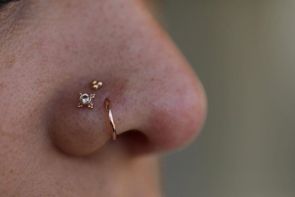 Nose with three gold piercings including a gem stud, a bead, and a hoop.