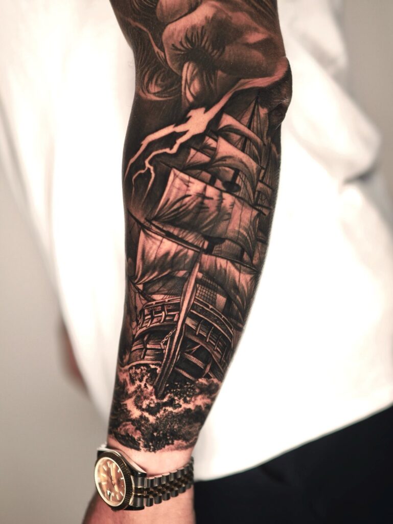 Detailed black and grey tattoo of a sailing ship on a person's forearm.